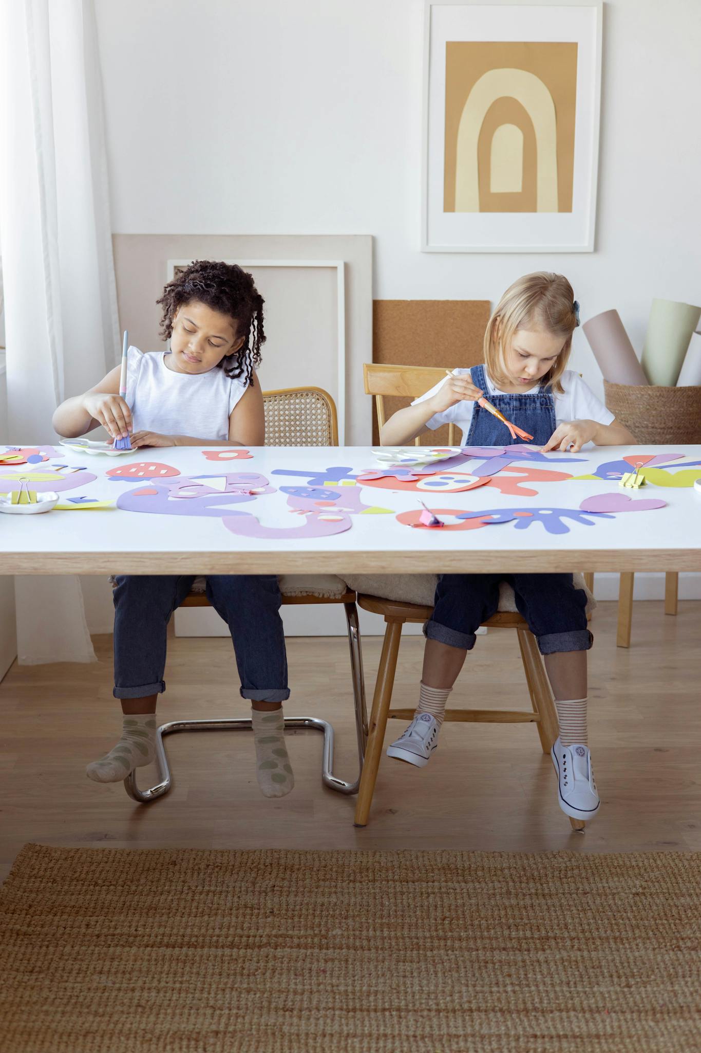 Two children enjoying arts and crafts, painting and cutting paper indoors at a table.